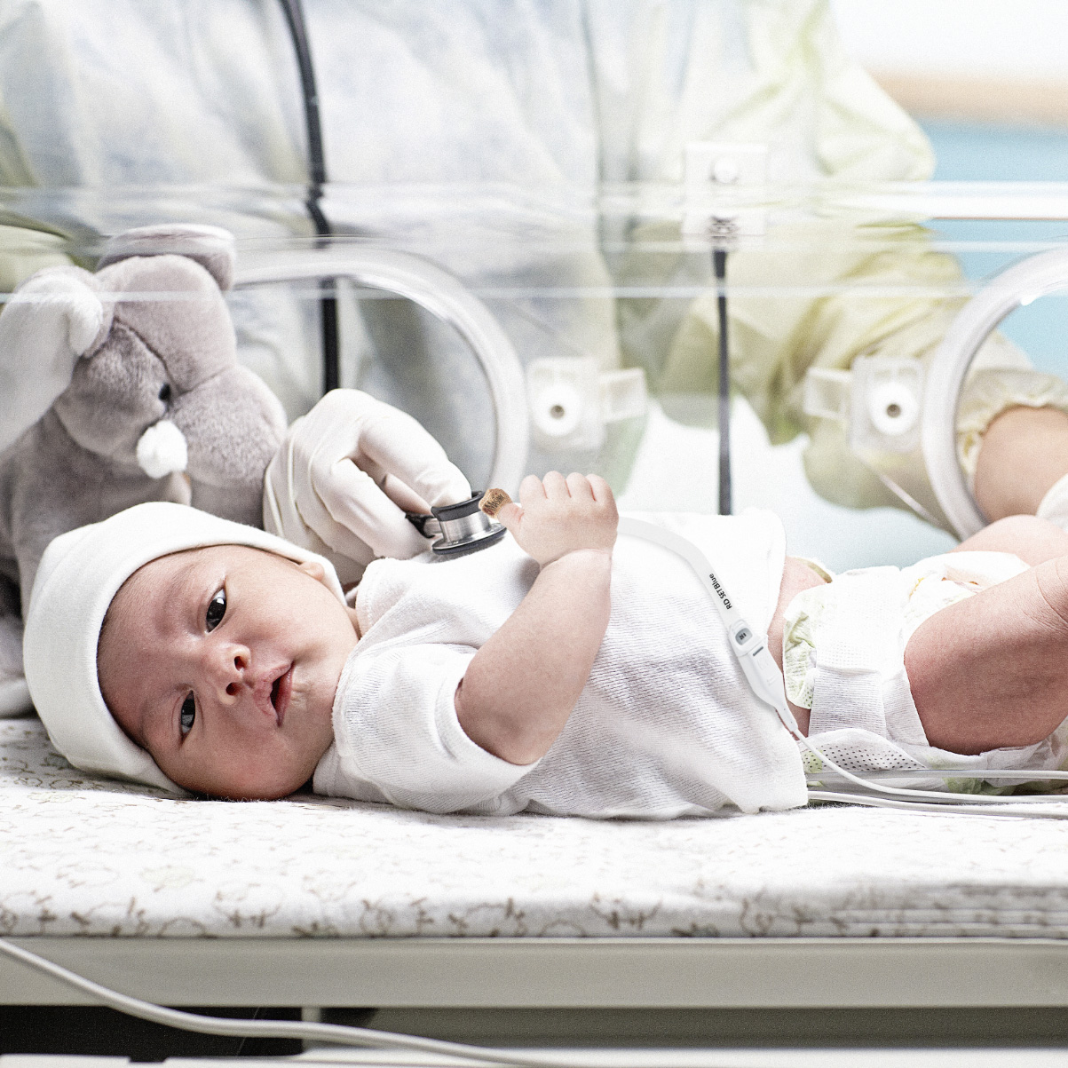 Infant in incubator in neonatal intensive care unit being examined by a clinician with a stethoscope and monitored with a Masimo RD SET pulse oximetry sensor.